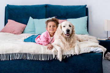 Curly african american kid lying on the bed with her golden retriever