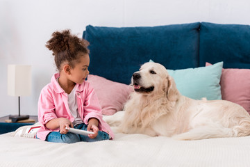 cute african american child sitting on the bed with golden retriever and playing with smartphone