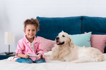 smiling african american kid sitting on the bed with smartphone and golden retriever