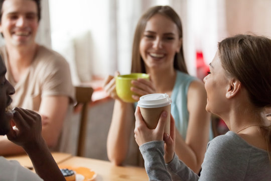Happy laughing diverse multiracial friends sitting at table drinking hot beverages, close up focus on cheerful caucasian girl holds coffee. Students spends free time together joking having fun in cafe