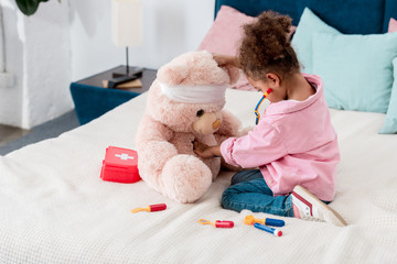 Little african american child in pink jacket playing the doctor  with teddy bear