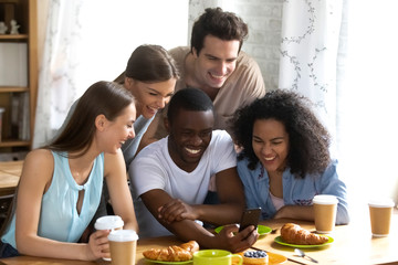 Cheerful diverse multiracial millennial people sitting at table indoors watching funny entertainment videos. Black guy met with best friends having fun spends free time together taking a selfie photo