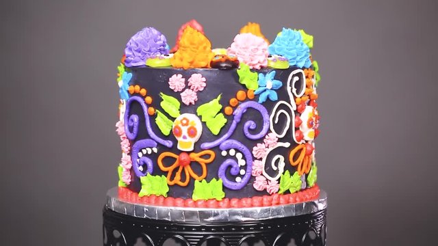 Gourmet Dia de los Muertos cake decorated with colorful buttercream frosting and gummy cupcake toppers on a black cake stand.