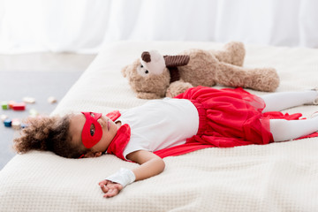 Cute little african american child in superhero costume and mask lying on bed with teddy bear