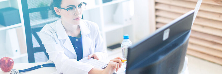 A beautiful young girl in a white robe is sitting at a computer desk with documents and a pen in...