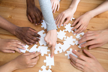 Close up diverse multiracial people hands assembling puzzle scattered on wooden table, top above view. Symbol and metaphor of teamwork and connection, business strategy and logic thinking concept