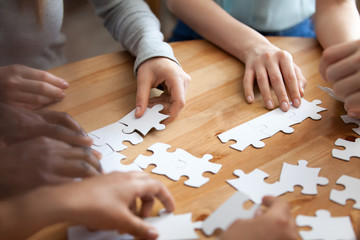 Close up diverse male female hands multiracial people assembling puzzle on wooden table cogitating making decision together. Team building and teamwork, business strategy and logic thinking concept