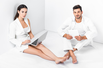 Obraz premium adult couple in total white sitting on floor and using digital devices