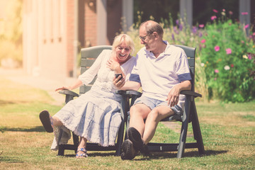 Happy retired couple sitting in the sunny garden, laughing together, looking on smartphone