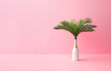 Champagne bottle with tropical green palm leaves on pastel pink background. Summer holiday concept with creative copy space. Summer tropical celebration party.