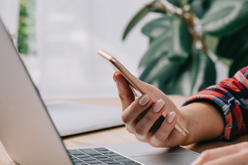 cropped shot of businesswoman with smartphone at workplace with laptop