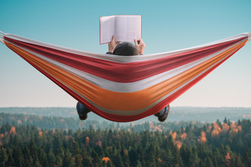 A man sits in a hammock and reads a book in a picturesque place. Back view. - obrazy, fototapety, plakaty