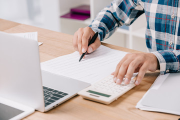 partial view of businessman making calculations at workplace with documents and laptop