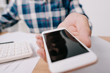 cropped shot of businessman showing smartphone with blank screen at workplace with papers and calculator