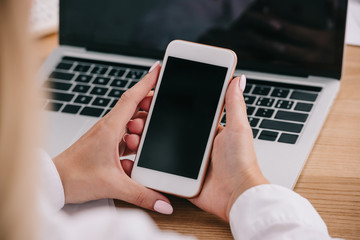 cropped shot of businesswoman holding smartphone at workpalce with laptop