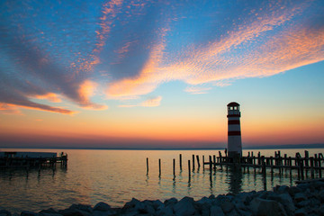 Sunset sky over the sea horizon landscape with lighthouse landmark with wooden dock pillars standing in the water, twilight scenic view from pier of Podersdorf am See, Neusiedl lake in Austria