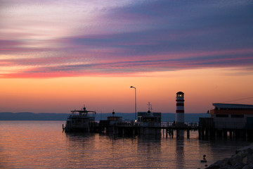 Obraz premium Beautiful twilight sky after sunset over the sea water with view of docked boats on pier and lighthouse tower with shadow reflection on the water, orange, yellow and violet colours
