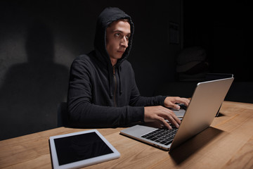 hacker in black hoodie using laptops at tabletop with tablet in dark room, cyber security concept