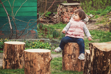 A little girl in a warm waistcoat and jeans sits on a stump in the village, ecotourism, holidays...