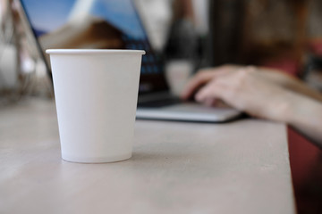 White Paper Cup of Coffee and Woman working as a Laptop on the background. Cup on workplace. Mockup. Freelance, office concept