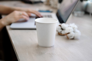White Paper Cup of Coffee and Woman working as a Laptop on the background. Cup on workplace. Mockup. Freelance, office concept