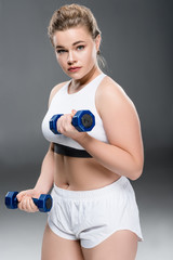 young size plus woman exercising with dumbbells and looking at camera on grey