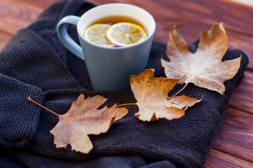 Autumn layout with hot tea with a lemon on a sweater and dry leaves. Brown background.
