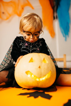 Portrait of little girl with painted face and fancy dress sitting on table with Jack O'Lantern pouting mouth