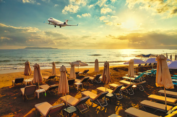 private business plane on landing flies over the sandy beach with sun loungers on the background of sunset, sun and clouds. Crete, Europe