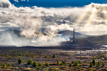 Environmental disaster. Copper plant in valley against the background of the mountains, toxic smoke from the pipes, dramatic rain clouds cut through the rays of the sun.