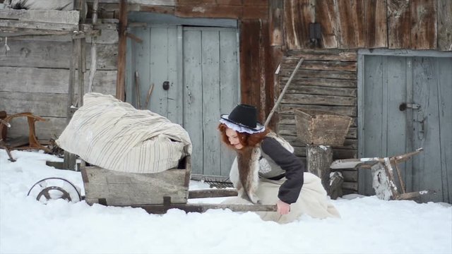 Charming red-haired girl carries a heavy cart in the winter near the old house. Hardworking Cinderella
