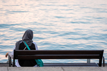 A woman with a scarf sits on a bench on the edge of water. View from the back.