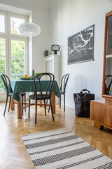 Real photo of white dining room interior with map poster, glass lamp, wooden table with fresh lemons and black chairs and rug on herringbone parquet