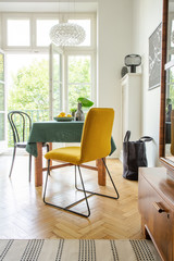 Mustard chair placed by the dining table with dark green tablecloth and fresh lemons in real photo of bright room interior with balcony and wooden floor