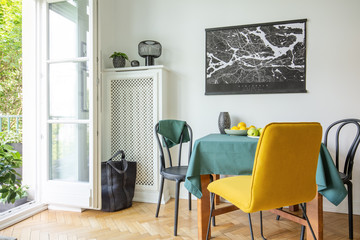 Map poster hanging on white wall in real photo of dining room interior with balcony, herringbone parquet, table with fruits and tablecloth with black and yellow chairs