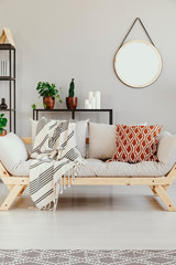 Blanket and cushion on wooden couch in ethno grey apartment interior with round mirror. Real photo