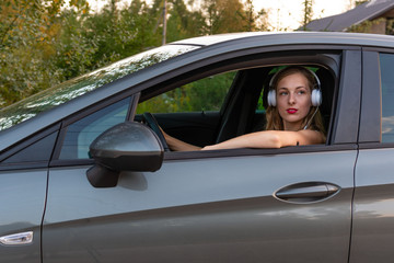 A young, beautiful woman with long hair and headphones sits at the wheel of the car.