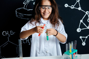 Mixed race asian, african girl in protective glasses wearing white lab holding test tubes and flasks conducting experiments over black board with symbols.
