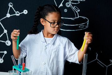 Mixed race asian, african girl in protective glasses wearing white lab holding test tubes and flasks conducting experiments over black board with symbols.