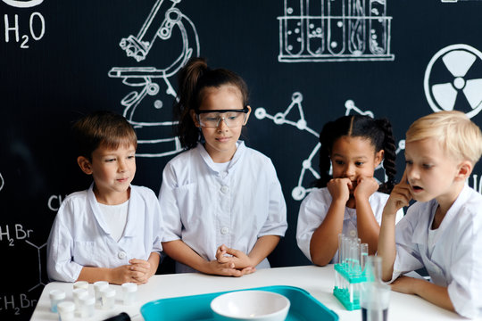 Surprised, shoked preteen multiracial schoolboys and girls in safety goggles, open-mouthed and bug eyed, leaning closely to the table and watching chemical reaction going on in the bowl