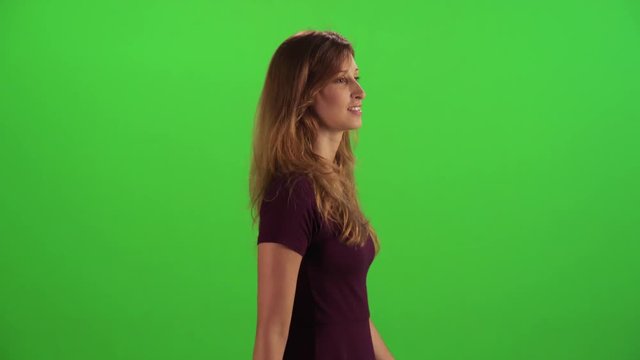 Young woman walking in a medium sideways shot over a green screen, looking around and to the back and waving hello.