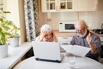 Happy cheerful old couple getting good news on email about their bank deposit, checking their annual interest income according papers in hands, man being in good mood shows thumb up