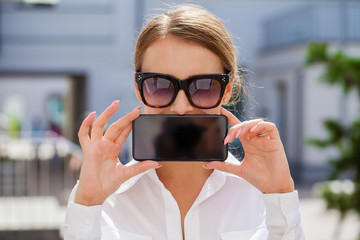 Young blonde woman showing your smartphone screen, summer park outdoors