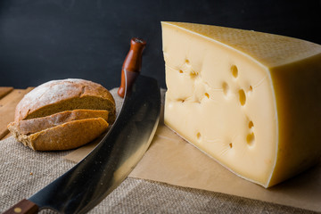 Composition of big piece of cheese and big two-handed knife on a dark background