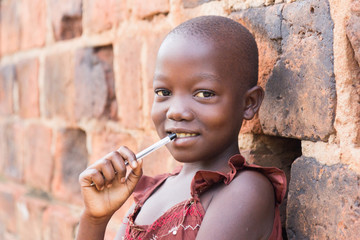 An 11-year old Ugandan girl smiling, holding a pen against her mouth and leaning against a brick...