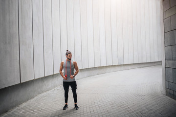 Strong and powerful athletist stands in grey corridor and looks to right. He is calm and peaceful. Guy has rest. He holds orange rope around his neck.