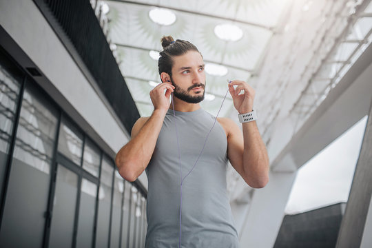 Picture of well-built and handsom bearded young man looks to right. He puts headphones in ears. Guy walks on stadium. He is alone.