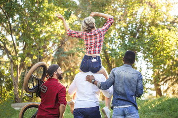 Rear view of unrecognized multiracial people going at the park in summer afternoon. Boyfriend carrying his girlfriend on piggyback and one of guys carries bmx byke over his shoulder.