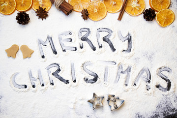 Christmas cooking: inscription merry christmas made from flour on a dark table, ingredients for baking and dried fruits on dark background