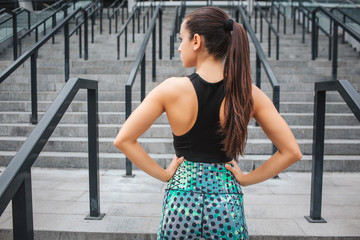 Calm and peaceful model stands and poses on cmera. She shows her back and buttocks. Young woman keeps ahnds on hips and looks to left. She is strong and well-built.